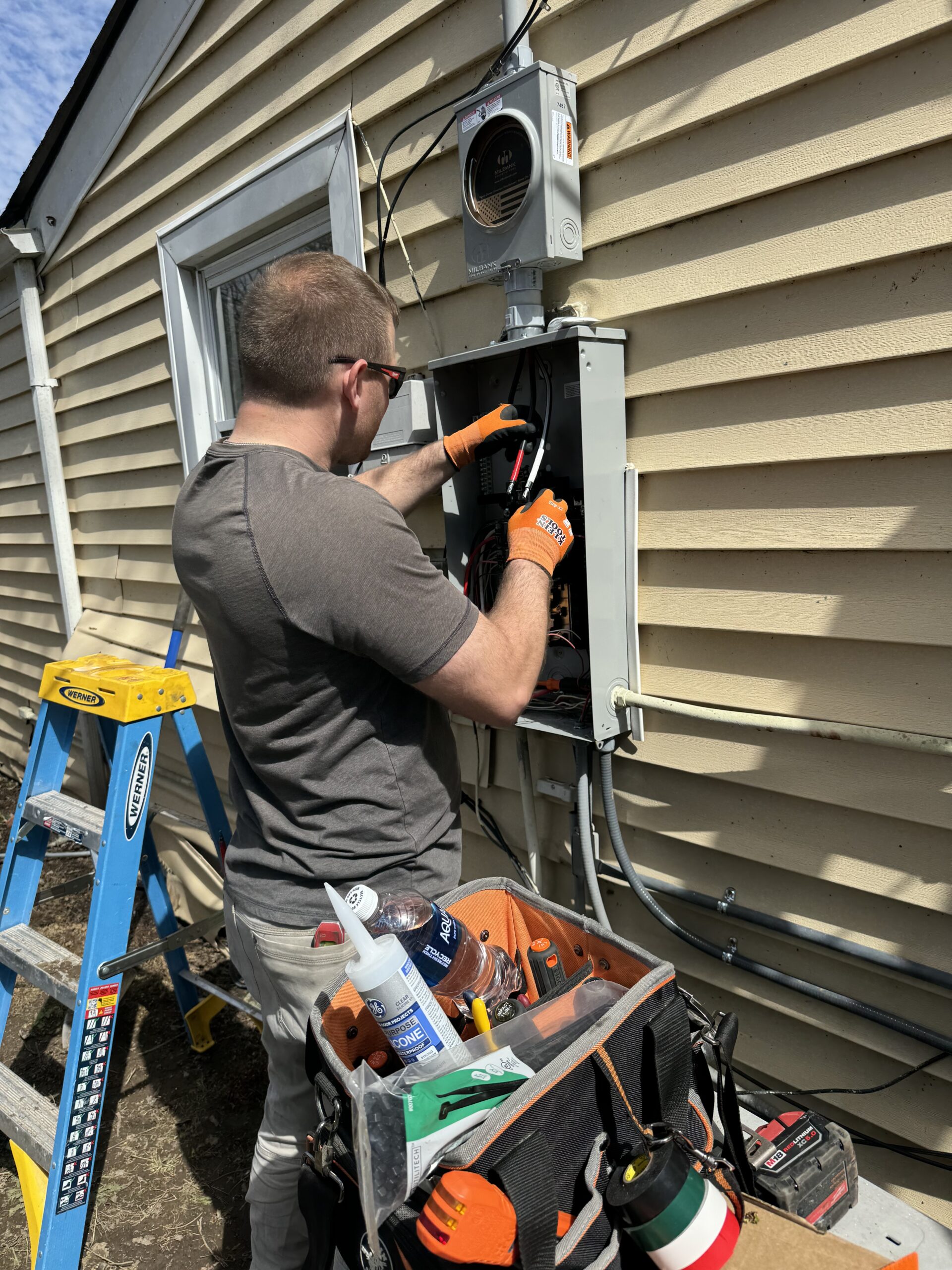 A man wearing gloves works on an outdoor electrical panel attached to a house, with tools, a ladder, and equipment nearby in the sunlight.