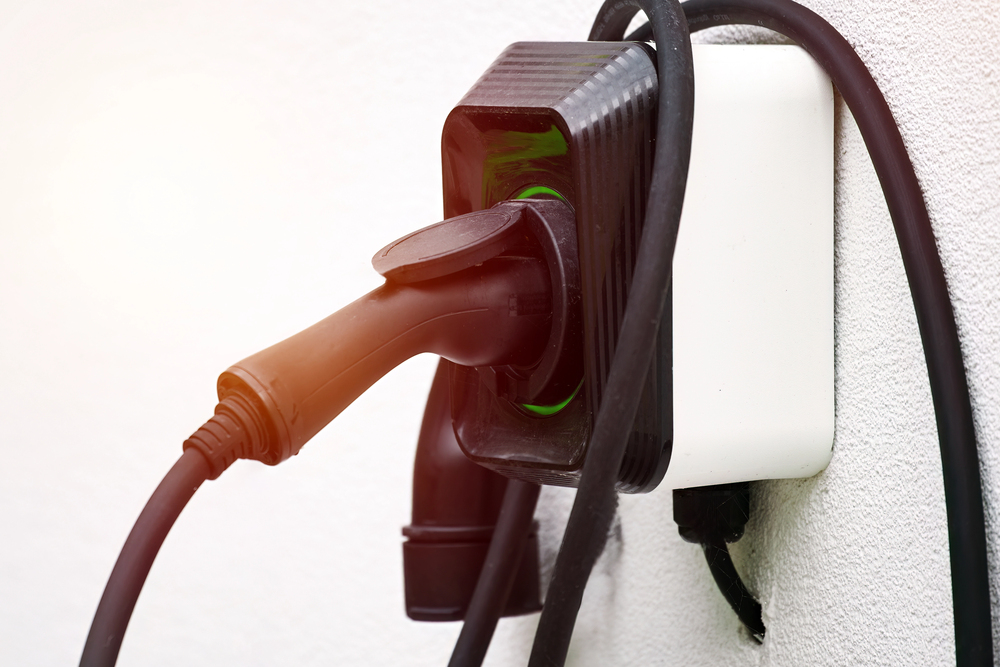 Close-up of a black electric vehicle charging plug inserted into a wall-mounted charging socket, with cables looped around the unit.