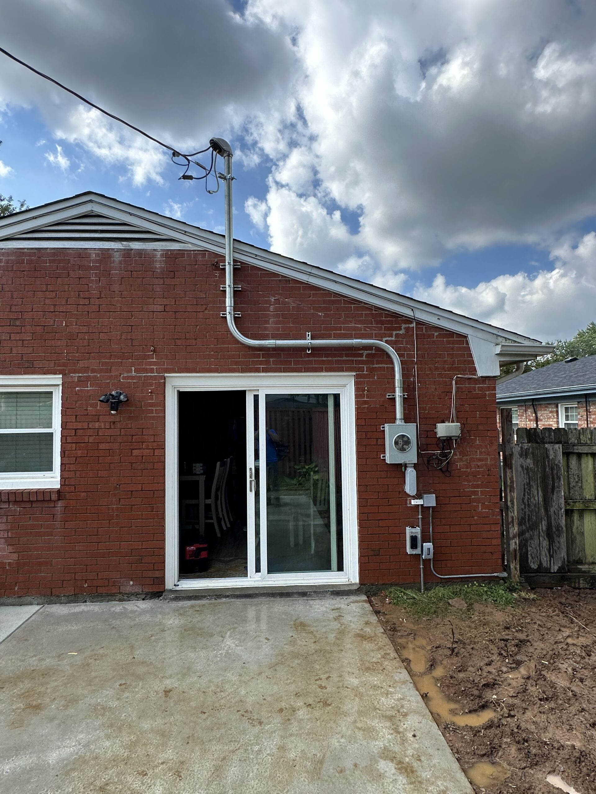 A red brick house with a white sliding glass door, outdoor electrical equipment and conduit, a wooden fence, and a cloudy sky overhead.