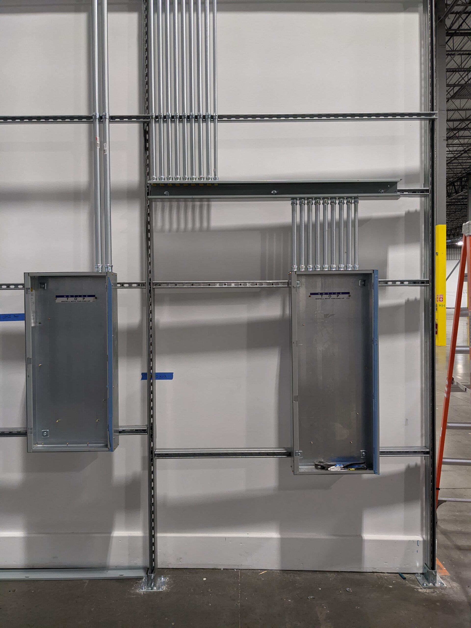 Two empty electrical panels mounted on a metal-framed wall with multiple conduit pipes running vertically and horizontally above and between them.