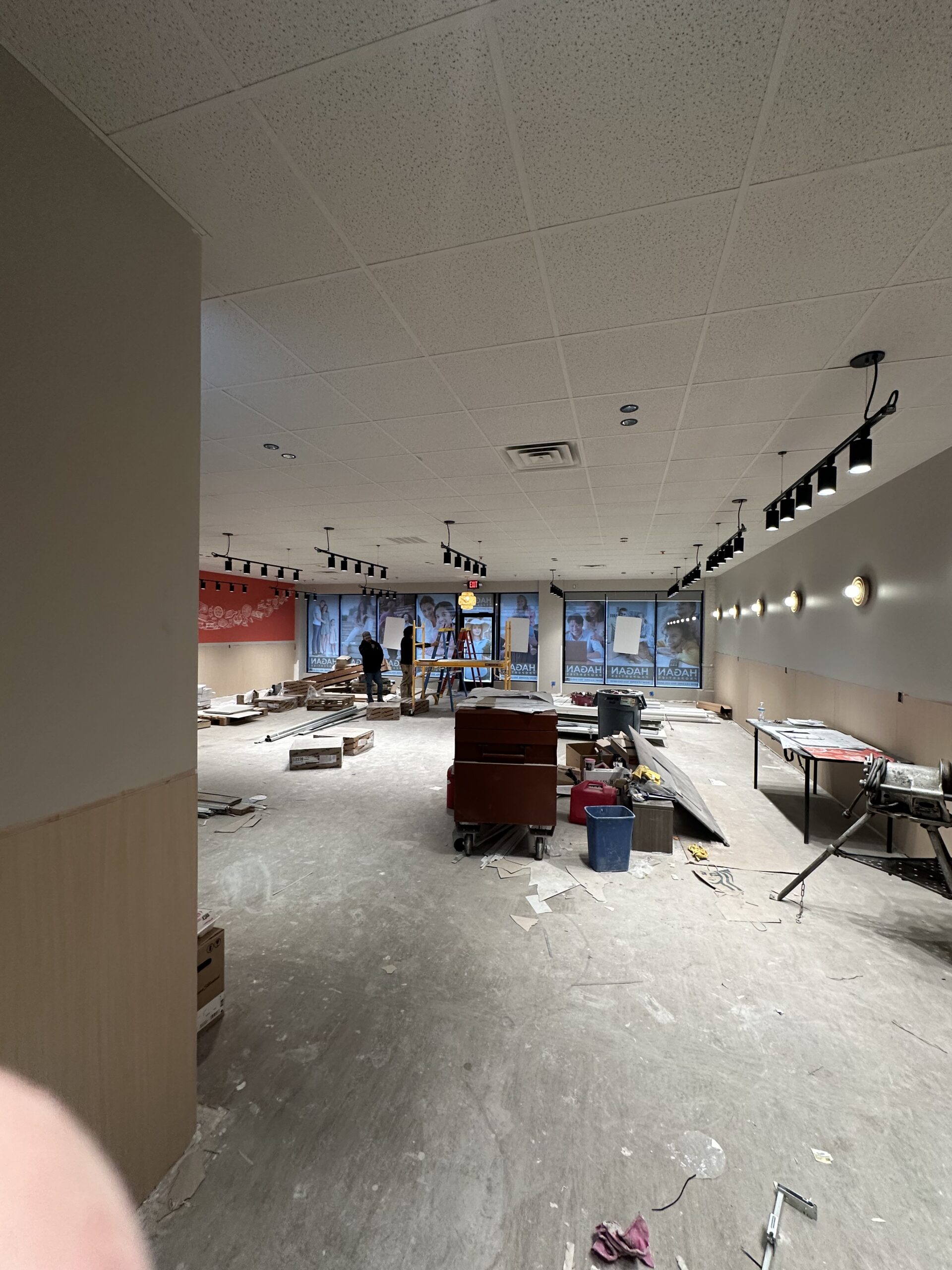 Interior of a commercial space under construction with scattered tools, building materials, and workers near the windows at the far end.