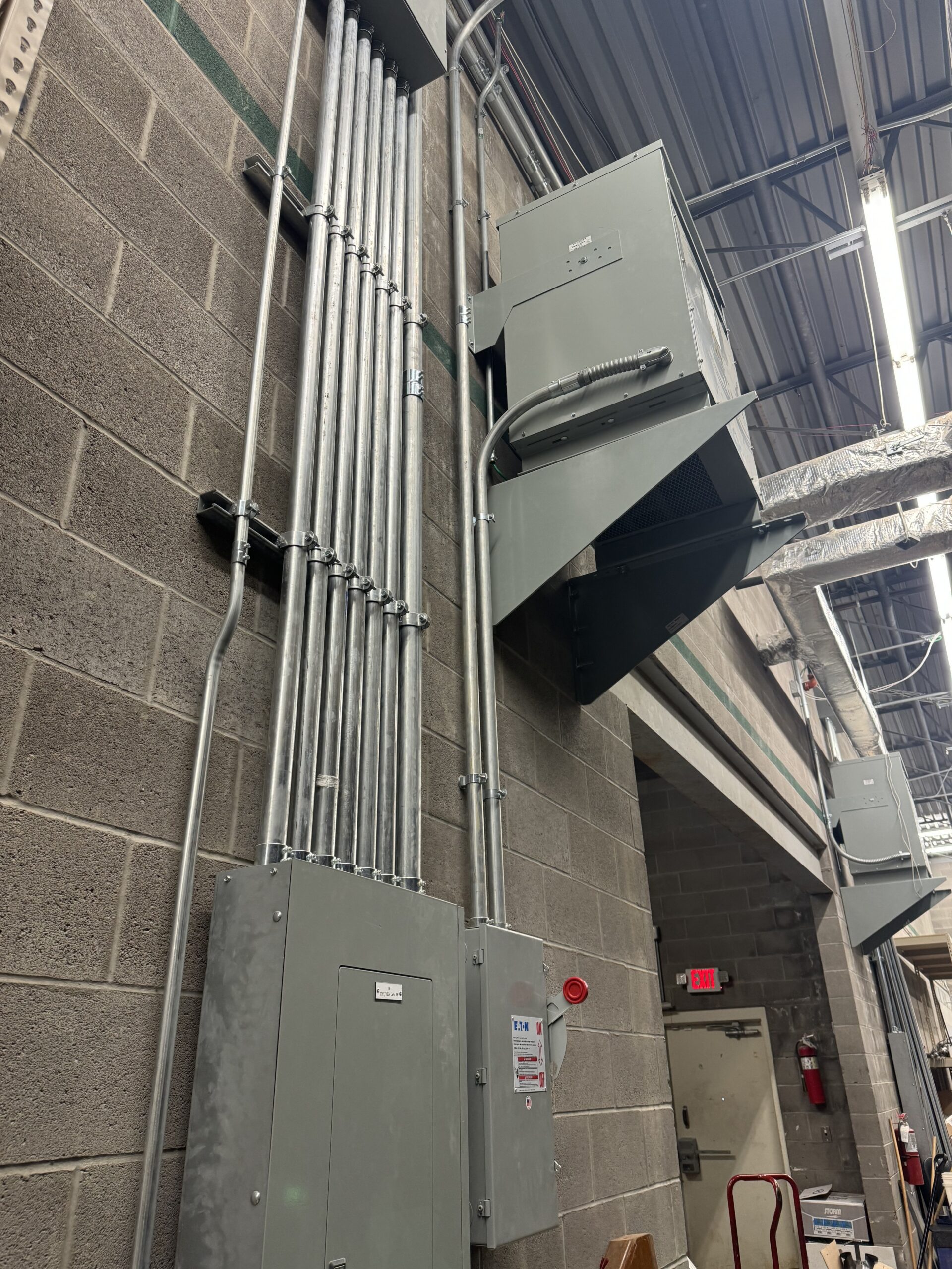 Several electrical conduit pipes and panels mounted on a concrete block wall inside a building showcase the quality of commercial electrical services in an industrial space with exposed ceiling and emergency exit.