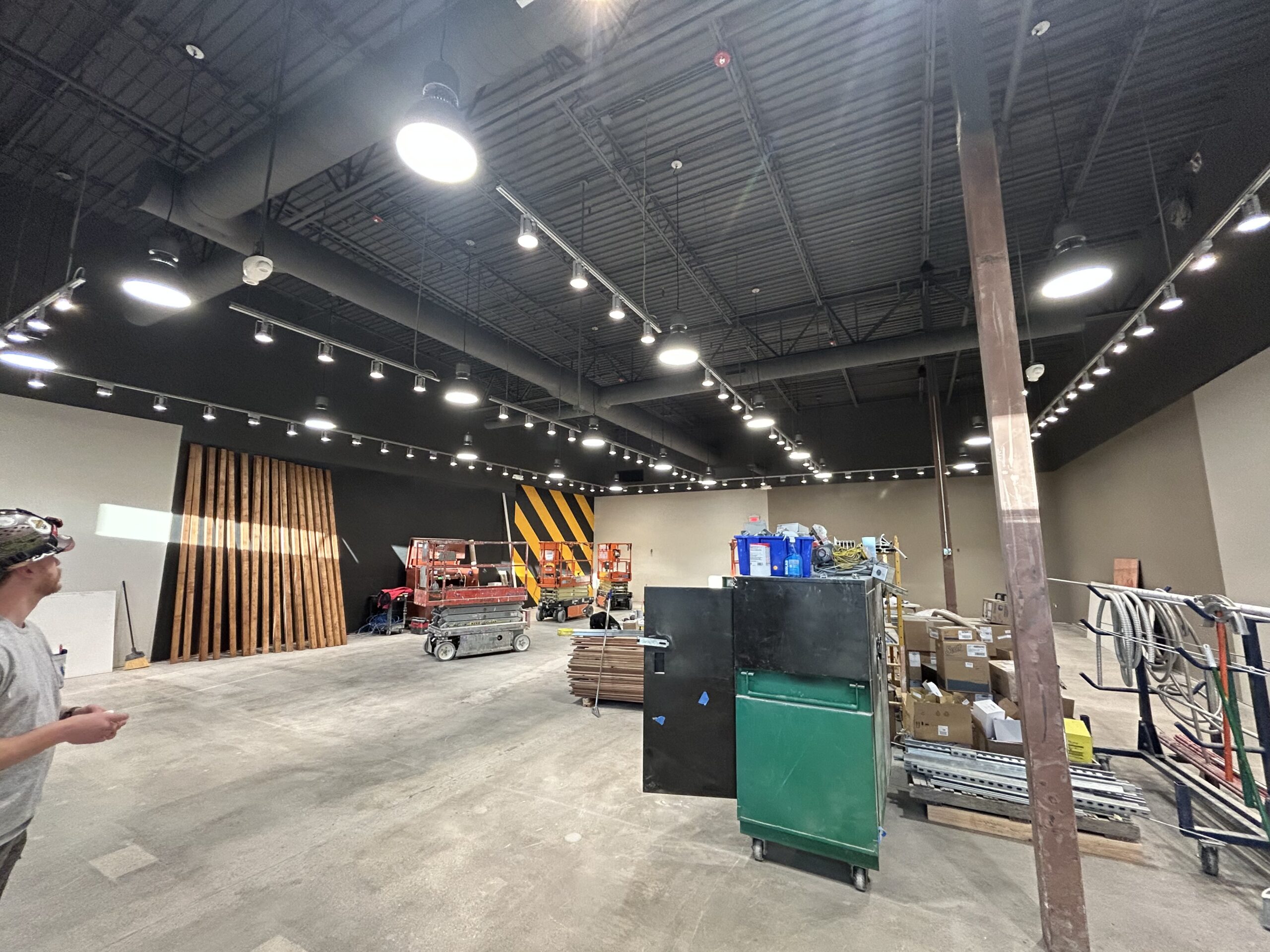 A large, unfinished commercial space with construction equipment, materials, and a person standing on the left side. Ceiling lights installed as part of commercial electrical services illuminate the area; walls are partially painted black with yellow stripes.