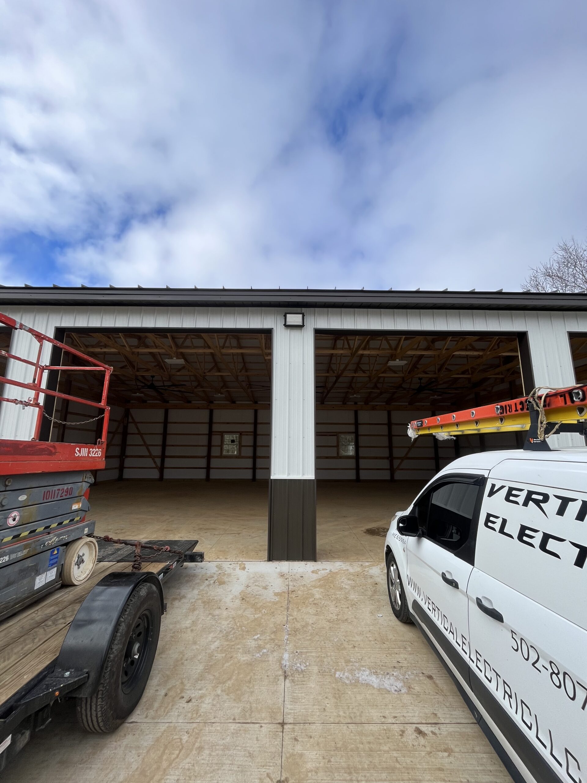 A white metal building with two large open garage bays, a red lift on a trailer to the left, and a white electrical service van parked on the right.